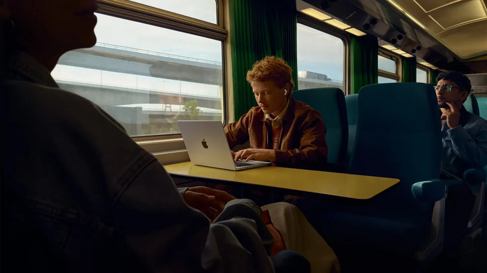 列車のテーブル席でノートパソコンを開き、AirPods Pro 3を装着して作業する若者を写したApple公式の画像