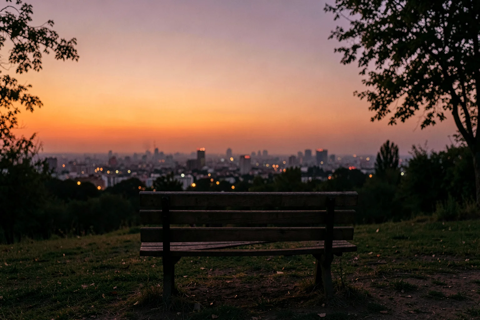 夕暮れの街並みを見下ろす高台のベンチと、オレンジ色に染まる空の風景